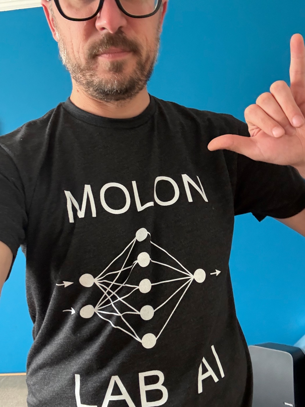 Ver: beard, heavy black glasses, pointing at his own t-shirt, which reads MOLON LAB AI around a neural-network diagram
